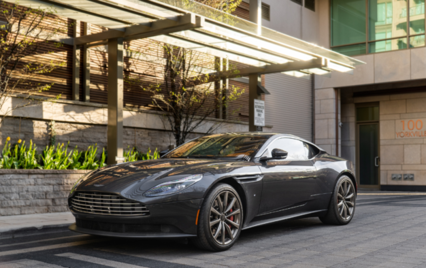 A dark gray Aston Martin sports car parked under a canopy in front of a building.