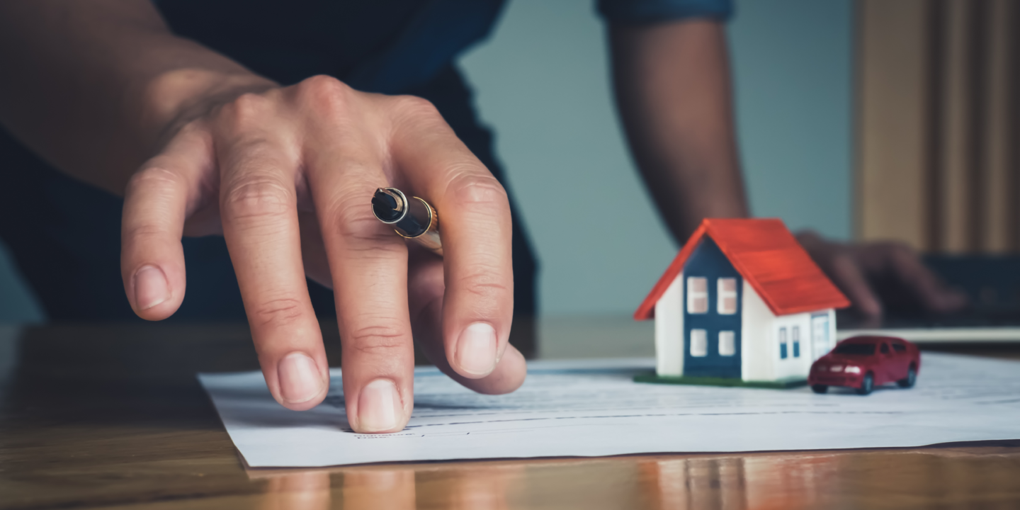 A person's hand holding a pen over a document, with a small model house and car in the background.