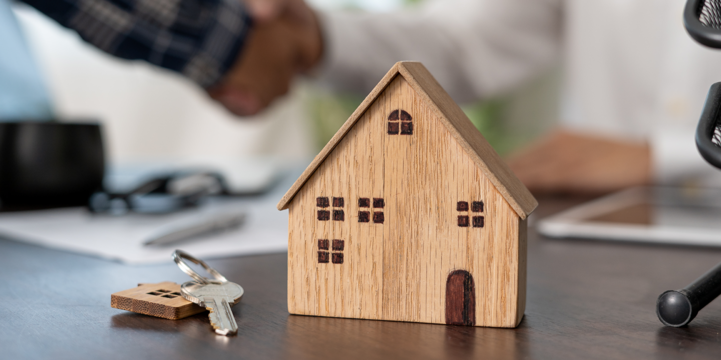 A wooden model house on a table with keys and a pen in the background.