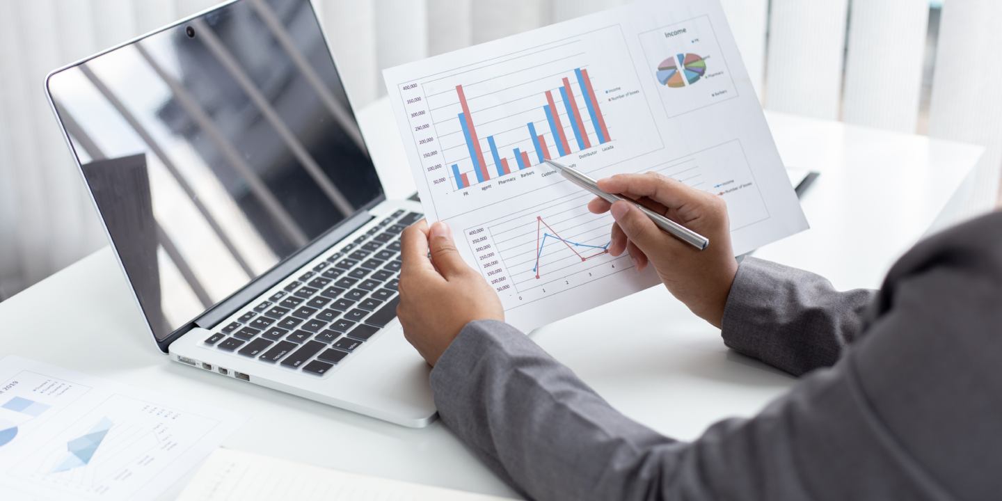 A person holding a printed report with bar charts, pie charts, and line graphs, sitting at a desk with an open laptop displaying a reflective screen.