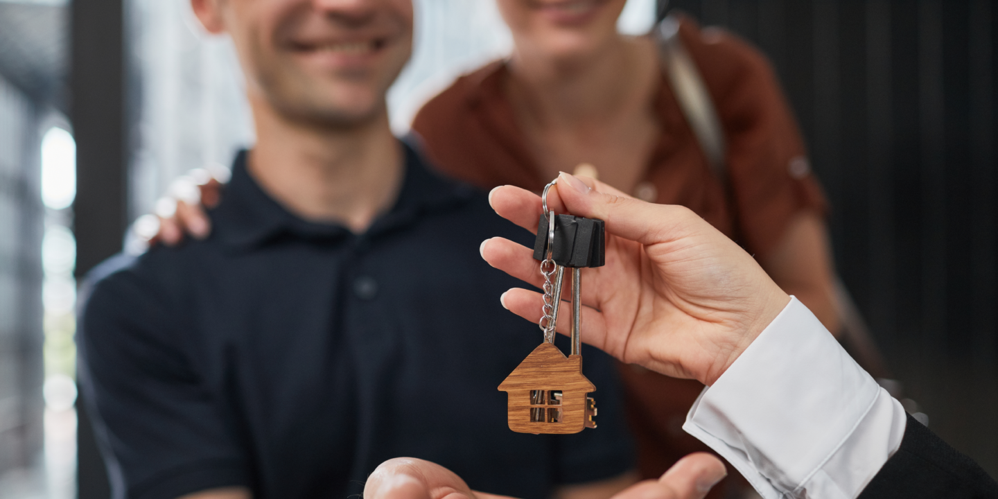 A man holds a house key, while a woman stands in the background, symbolizing homeownership and partnership.