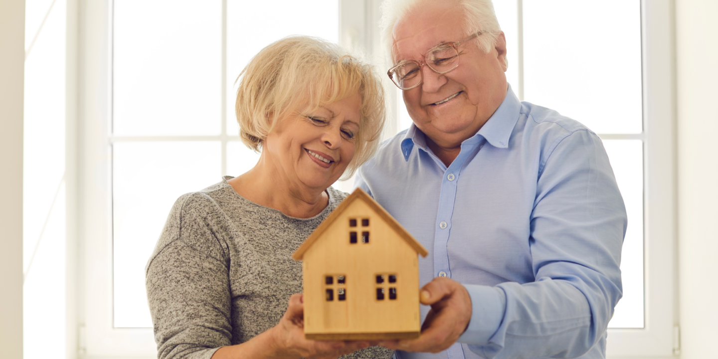 A senior couple proudly displays a wooden house model, representing their shared journey and aspirations for the future.