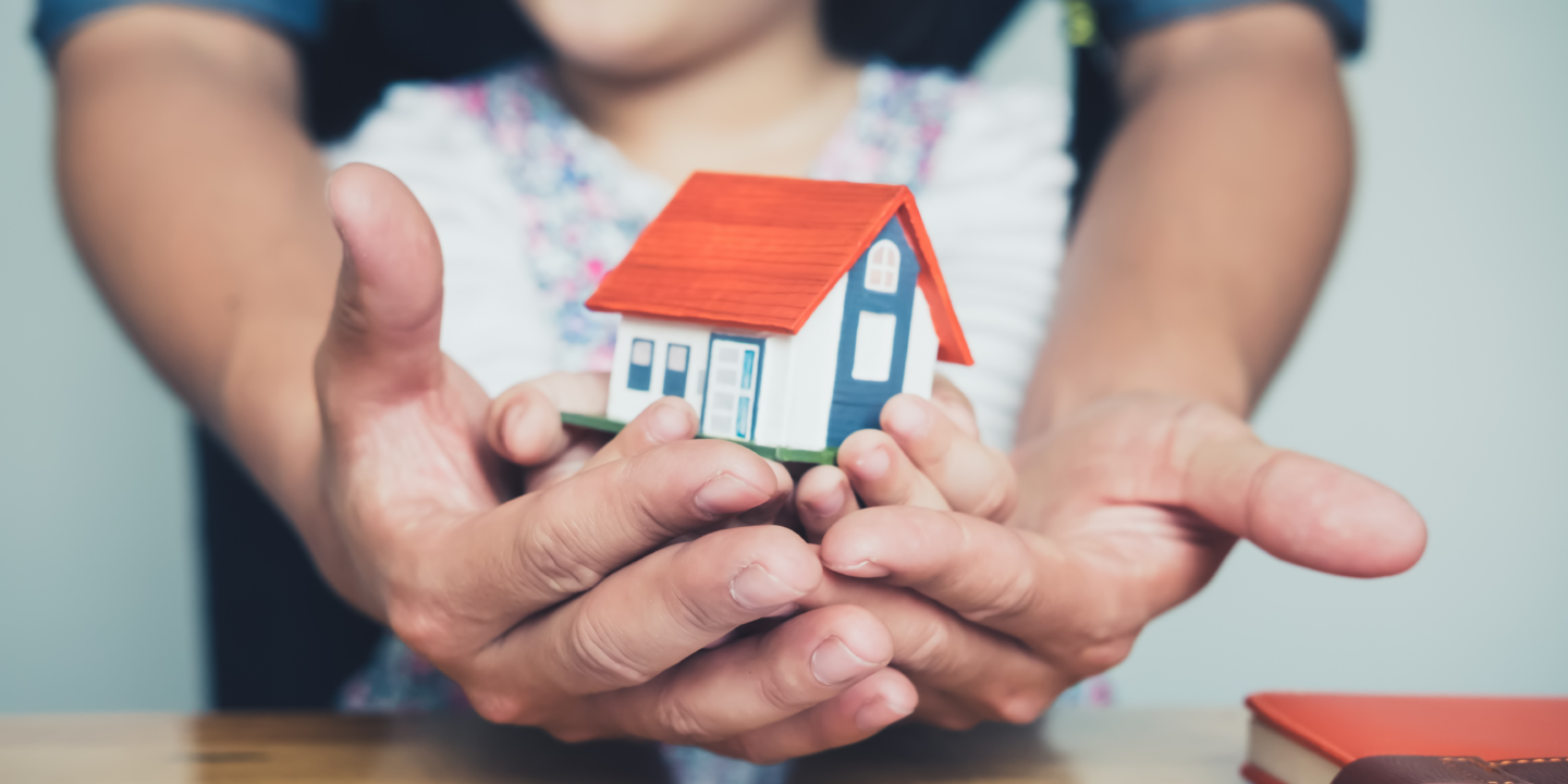 A person gently cradles a small house model in their hands, symbolizing care.