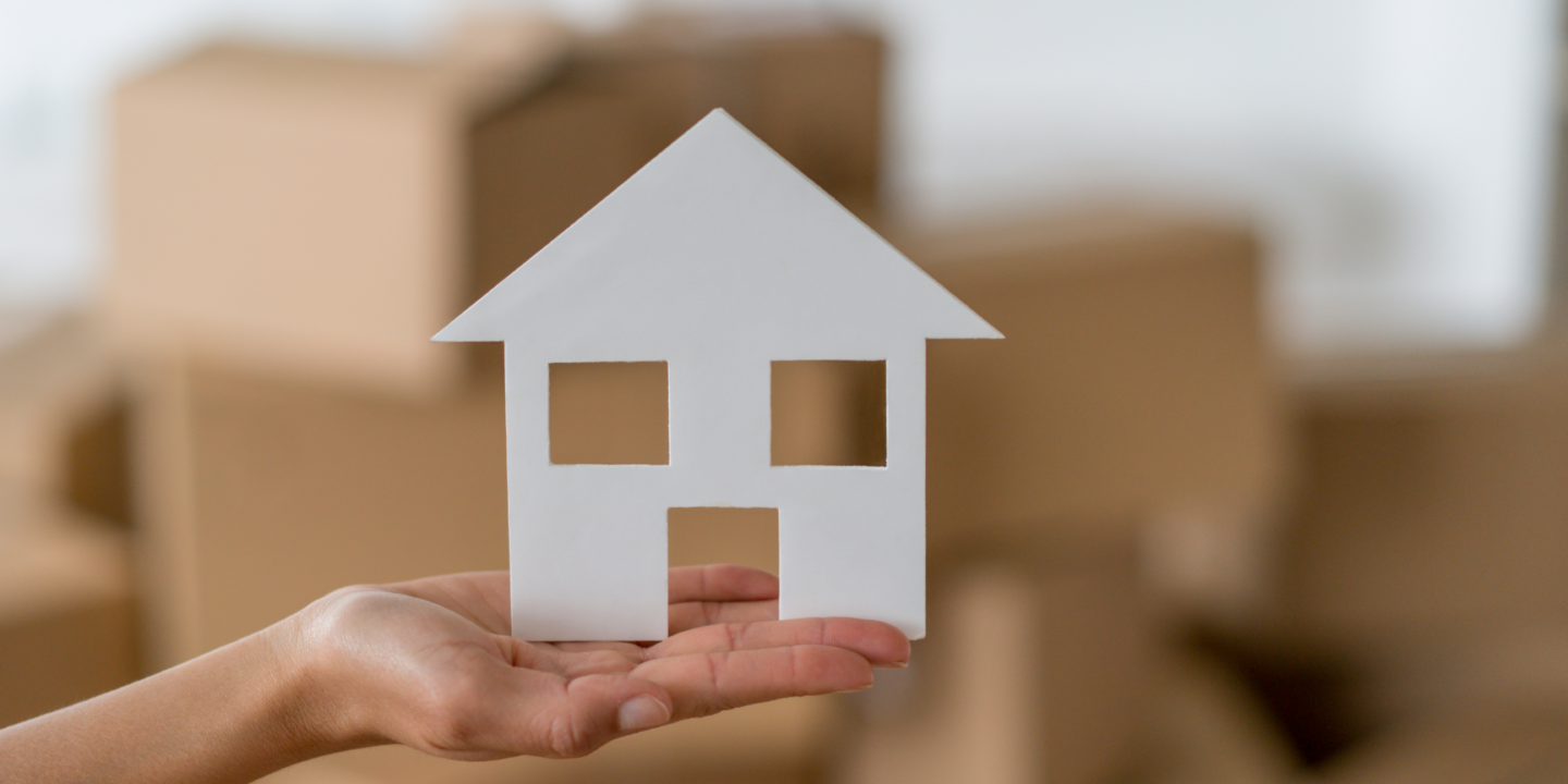 A person holds a paper house in front of a stack of moving boxes, symbolizing a new beginning or relocation.